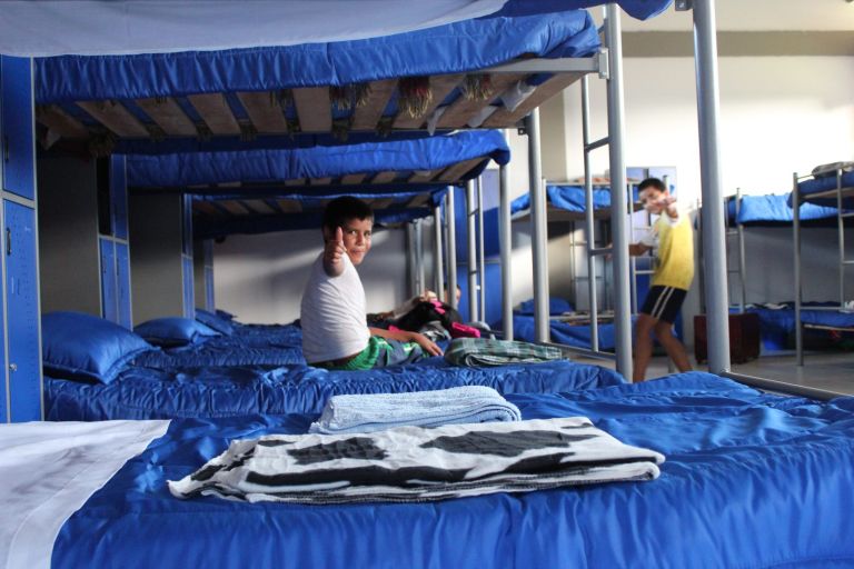 Un niño levanta el pulgar desde su camarote en el nuevo dormitorio, con cobijas azules, casilleros y pisos de baldosa.