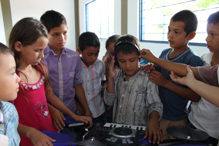 Un grupo de niños rodea a un compañero que prueba unos audífonos frente a un teclado de música en el aula nueva.