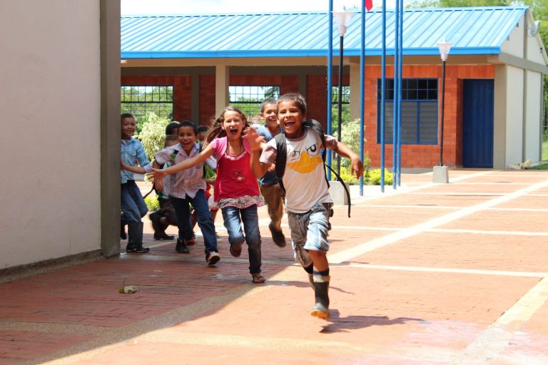 Niños corren riendo por la plazoleta de ladrillo, frente a las aulas de techo azul.