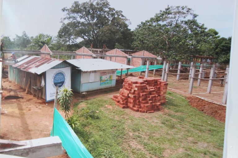 La obra en marcha: los nuevos bloques de ladrillo se levantan junto a las aulas de tablas del antiguo albergue, con una pila de ladrillos en primer plano.