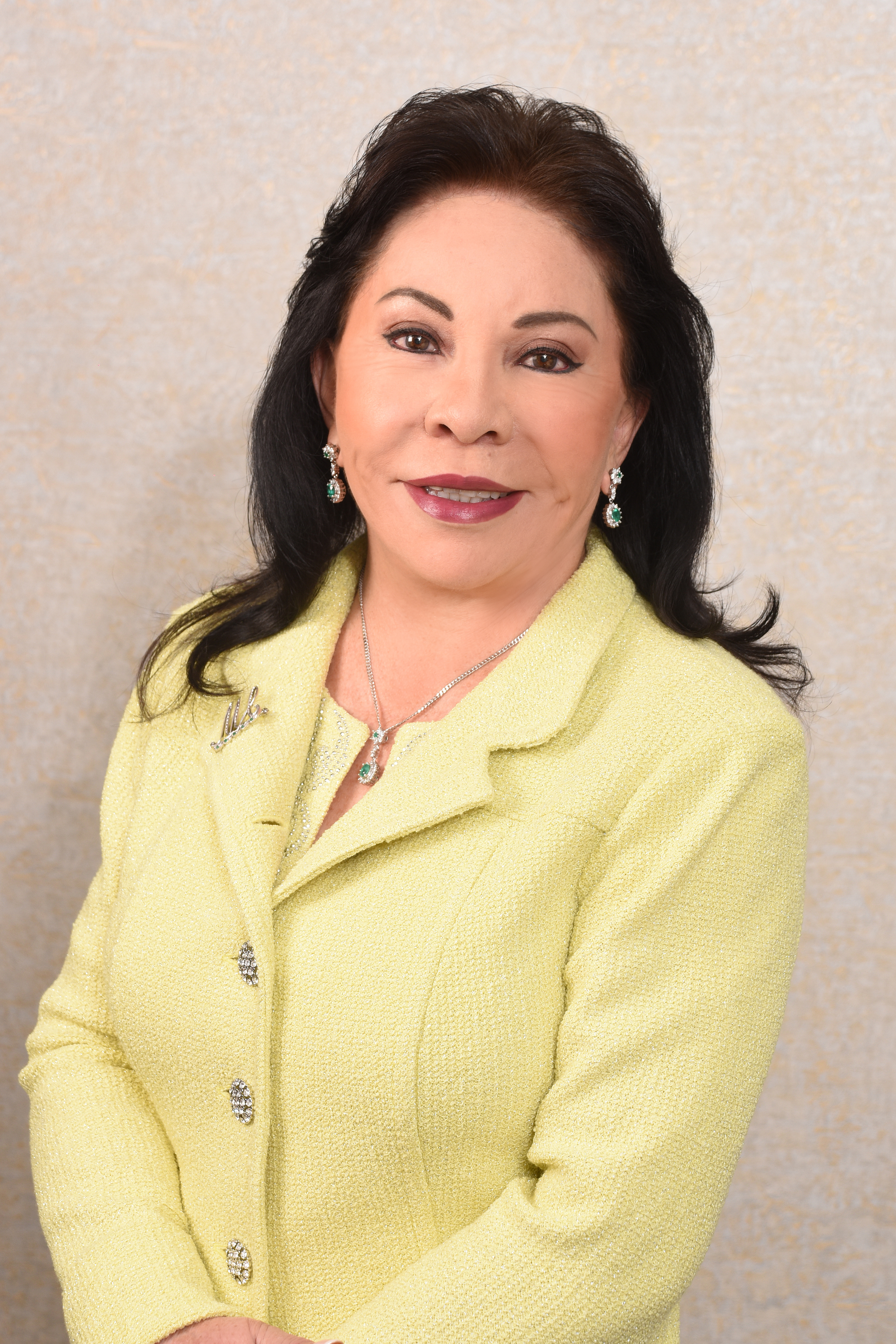 Foto de la Dra. Maria Luisa fundadora de la Fundación Internacional Maria Luisa de Moreno (FIMLM). Mirando sonriente a la cámara, vestida de manera formal con traje amarillo, con una pared de fondo en tono beige claro.