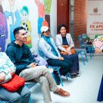 Personas esperando en una sala para ser atendidas durante un evento comunitario, mientras un voluntario mujer de la Fundación les explica procedimientos y demás.