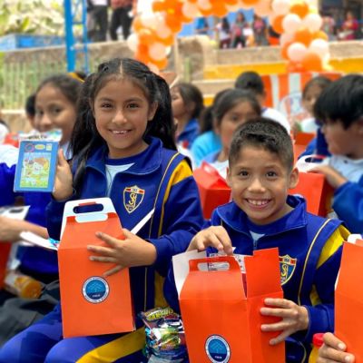 Una niña y un niño miran a la cámara sonríen mostrando sus regalos, una cajita con palomitas de maíz, un juego didáctico y el refresco para ver el Tele cine