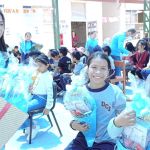 La voluntaria de la Fundación esta cerca de una niña y un niño con una gran caja de regalos que contienen los conos sorpresas para regalar y les entrega