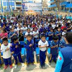 Mas de cien niños puestos de pie con la mano en el pecho cantan con alegría el himno nacional del Perú.