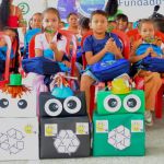 Niños sentados en sillas rojas sostienen figuras decorativas mientras posan junto a coloridas cajas disfrazadas como contenedores de reciclaje con ojos y sombreros, representando la separación de residuos en blanco, negro y verde.