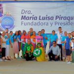 Grupo de voluntarios y organizadores posan frente a un gran cartel de la Dra. María Luisa Piraquive, en el marco de un evento educativo de jornada de apoyo sobre reciclaje y entrega de kits escolares.
