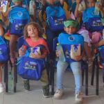 Un grupo de niños pequeños sentados en sillas sostienen con orgullo sus nuevas mochilas azules, botellas de agua y cajas de refrigerios, algunos llevando antifaces de superhéroe.