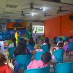Niños y niñas junto a sus acompañantes observan atentos una presentación sobre valores ambientales, mientras ecoheroe captan su atención con actividades lúdicas.